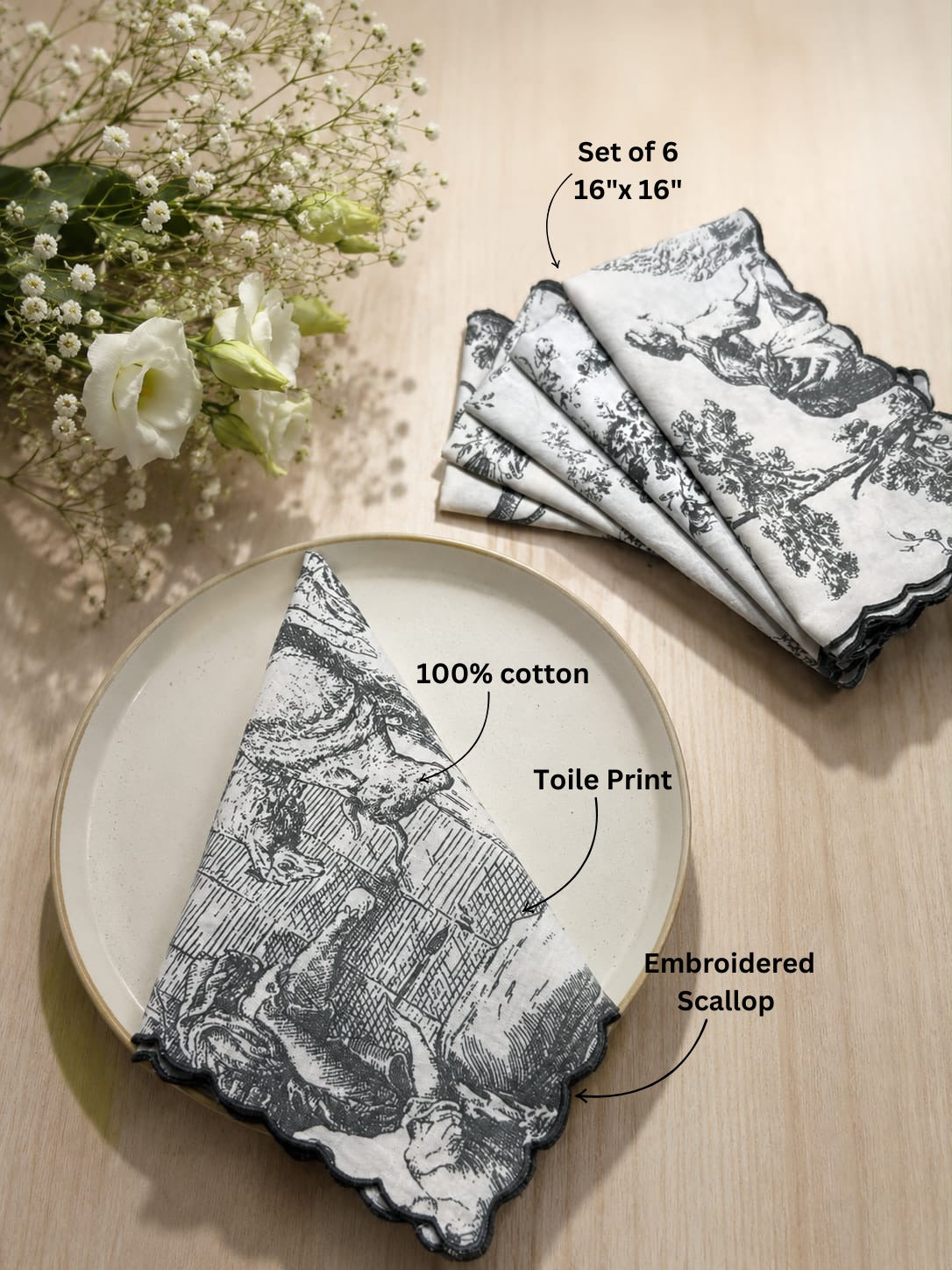 A set of six folded cotton napkins with a black and white toile print and scalloped edges, displayed on a light wood surface with decorative flowers. Text overlays highlight features like 'Set of 6 16"x16"', '100% cotton', 'Toile Print', and 'Embroidered Scallop'.