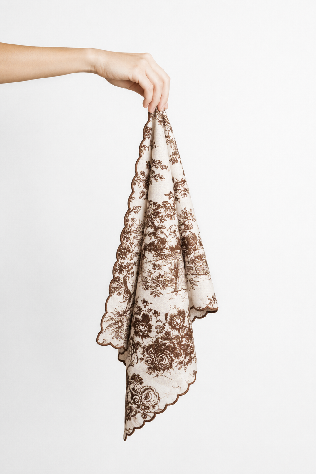 A hand holding a square napkin with a brown toile floral print on a cream background, featuring a scalloped edge, against a plain white background.