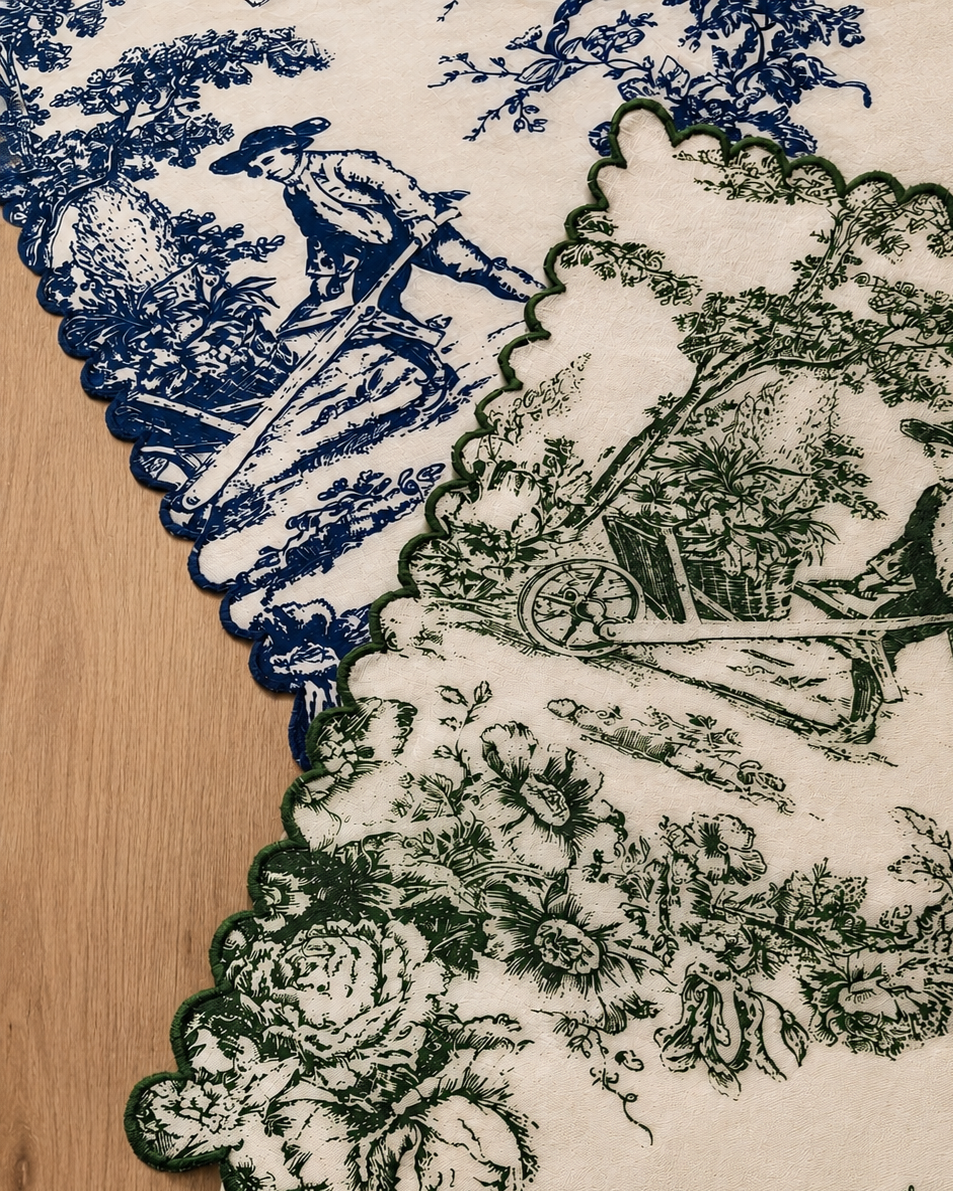 A close-up of a scalloped fabric or tablecloth with a toile-style print, showing blue and dark green illustrations on an off-white background, laid over a light wood surface.