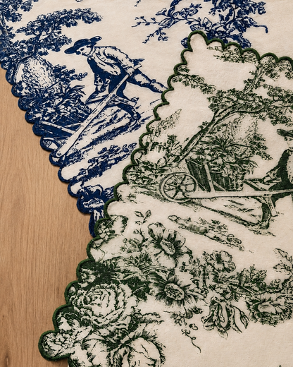 A close-up of a scalloped fabric or tablecloth with a toile-style print, showing blue and dark green illustrations on an off-white background, laid over a light wood surface.