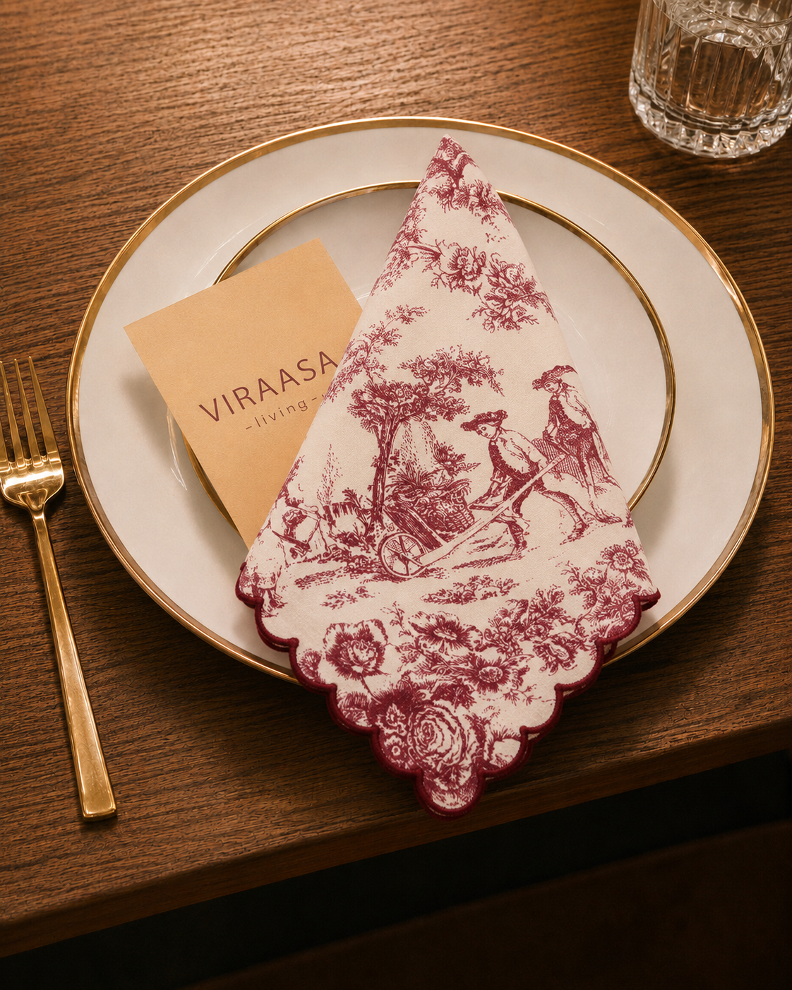 A folded cream napkin with a dusty rose toile de Jouy pattern is styled on a white plate with a gold rim, alongside a gold fork and a small Viraasa dining card, set on a dark wood table.